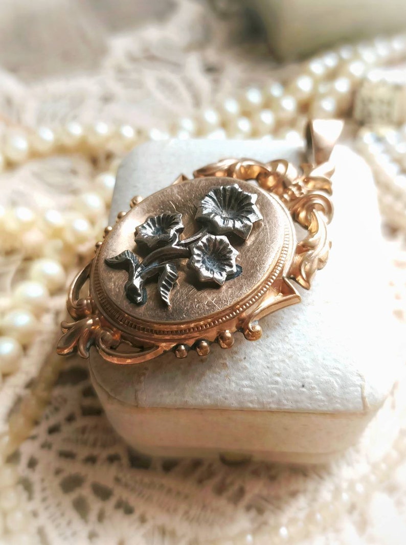 K&ouml;nnte beinhalten: Ein goldenes Medaillon mit einem floralen Design. Das Medaillon ist oval geformt und hat auf der Vorderseite ein zartes Blumenmuster. Die Blumen sind aus Silber und haben einen schwarzen Emaille-Mittelpunkt.