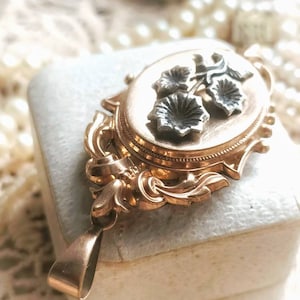 K&ouml;nnte beinhalten: Ein goldenes Medaillon mit floralem Design. Das Medaillon hat einen zarten, verzierten Rahmen und zeigt drei silberne Blumen mit einem Stiel.