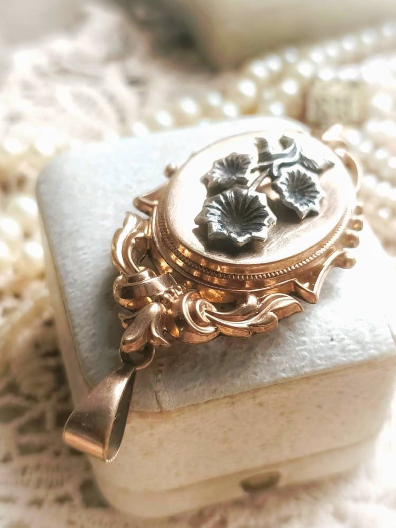 K&ouml;nnte beinhalten: Ein goldenes Medaillon mit floralem Design. Das Medaillon hat einen zarten, verzierten Rahmen und zeigt drei silberne Blumen mit einem Stiel.
