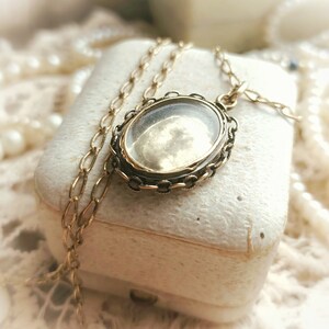 May include: A gold locket necklace with a clear glass oval pendant. The locket is on a white box with a pearl necklace in the background.