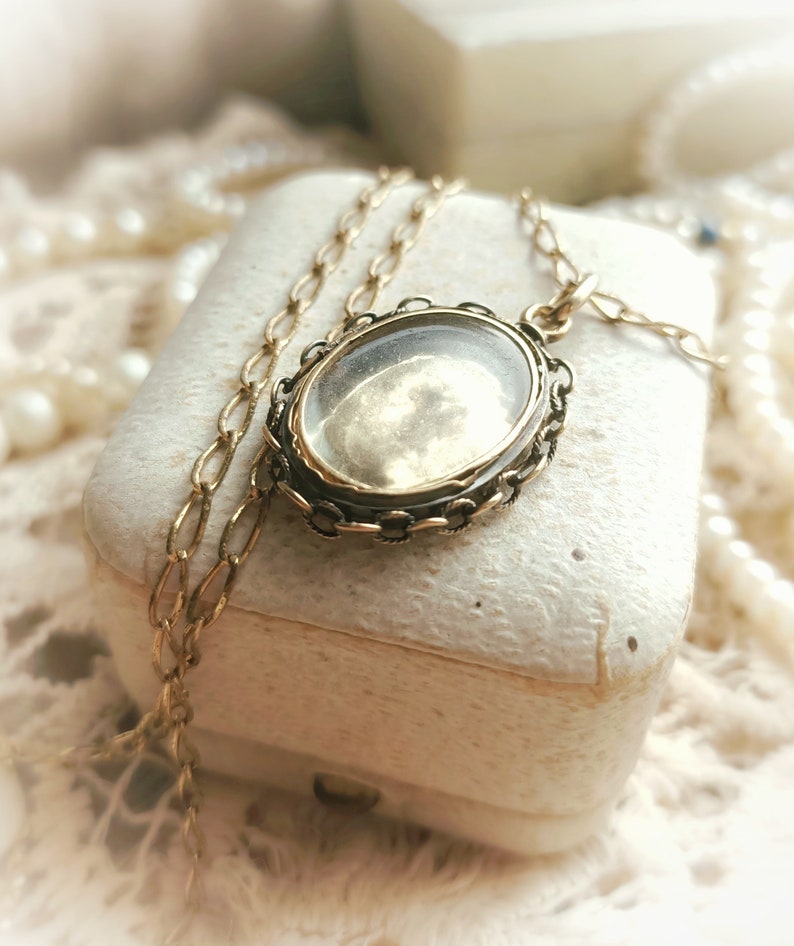 May include: A gold locket necklace with a clear glass oval pendant. The locket is on a white box with a pearl necklace in the background.