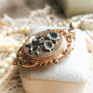 K&ouml;nnte beinhalten: Ein goldenes Medaillon mit einem floralen Design. Das Medaillon ist oval geformt und hat auf der Vorderseite ein zartes Blumenmuster. Die Blumen sind aus Silber und haben eine schwarze Emaille-Oberfl&auml;che.