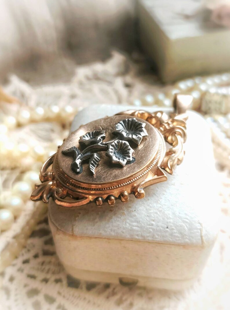 K&ouml;nnte beinhalten: Ein goldenes Medaillon mit einem floralen Design. Das Medaillon ist oval geformt und hat auf der Vorderseite ein zartes Blumenmuster. Die Blumen sind aus Silber und haben eine schwarze Emaille-Oberfl&auml;che.