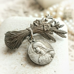 May include: A silver-toned brooch featuring a bouquet of wheat stalks and flowers with a round pendant featuring a profile of a woman with the text "JEANNE D'ARC" and "SAINTE" around the edge.