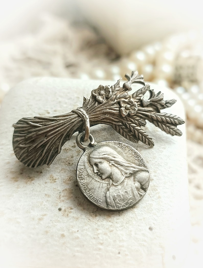 May include: A silver-toned brooch featuring a bouquet of wheat stalks and flowers with a round pendant featuring a profile of a woman with the text "JEANNE D'ARC" and "SAINTE" around the edge.