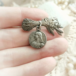 May include: Silver tone brooch featuring a bouquet of wheat and a round pendant with a depiction of a woman's face. The pendant has the text "Bienheureuse" and "Sainte" around the edge.