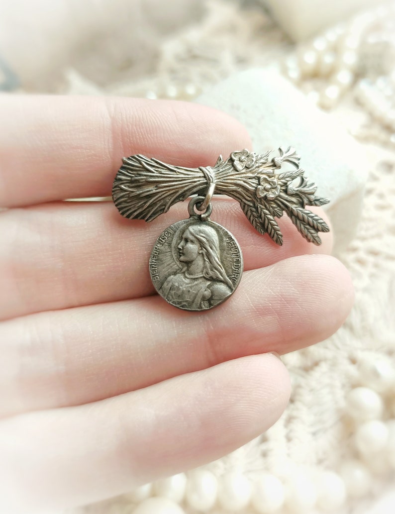 May include: Silver tone brooch featuring a bouquet of wheat and a round pendant with a depiction of a woman's face. The pendant has the text "Bienheureuse" and "Sainte" around the edge.