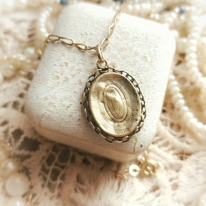 May include: A gold oval locket pendant with a detailed engraved design, hanging from a gold chain. The locket is resting on a white surface.