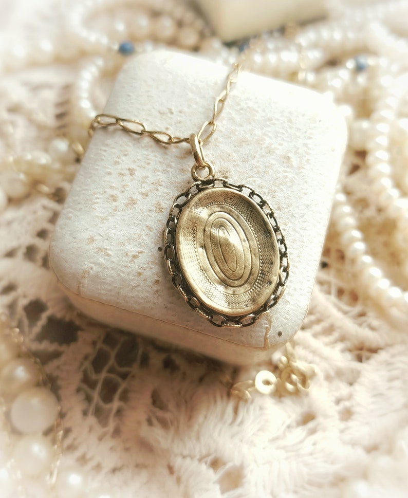 May include: A gold oval locket pendant with a detailed engraved design, hanging from a gold chain. The locket is resting on a white surface.