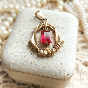 May include: A gold pendant with a red gemstone set in a geometric frame. The pendant is in a white box with a pearl necklace in the background.