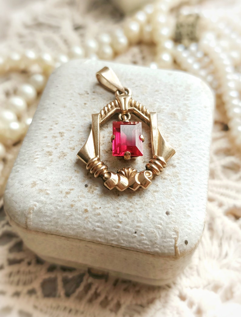 May include: A gold pendant with a red gemstone set in a geometric frame. The pendant is in a white box with a pearl necklace in the background.