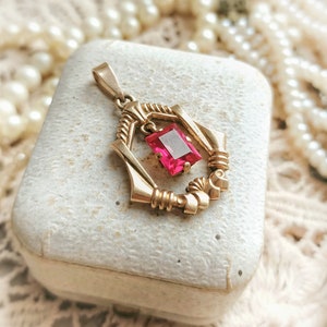 May include: A gold pendant with a pink gemstone set in a geometric design. The pendant is sitting on a white box.