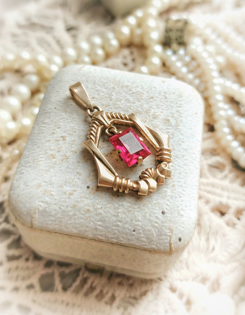 May include: A gold pendant with a pink gemstone set in a geometric design. The pendant is sitting on a white box.