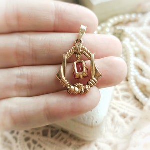 May include: A gold pendant with a red gemstone set in a geometric frame. The pendant is suspended from a gold bail.