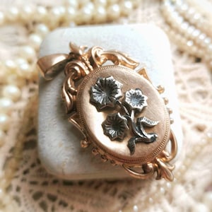 K&ouml;nnte beinhalten: Ein goldenes Medaillon mit einem floralen Design. Das Medaillon ist oval geformt und hat auf der Vorderseite ein detailliertes florales Design. Die Blumen sind aus Silber und auf einem goldenen Hintergrund angebracht.