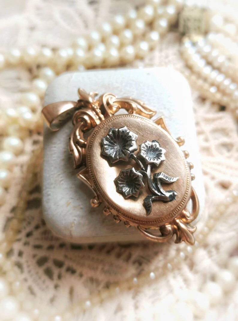K&ouml;nnte beinhalten: Ein goldenes Medaillon mit einem floralen Design. Das Medaillon ist oval geformt und hat auf der Vorderseite ein detailliertes florales Design. Die Blumen sind aus Silber und auf einem goldenen Hintergrund angebracht.