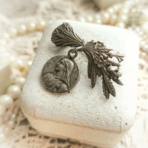May include: A silver pendant featuring a religious medal with the image of a woman and the text "BEAUFIGURISE" and "SACI MARIE". The pendant is attached to a silver charm shaped like a bouquet of wheat.