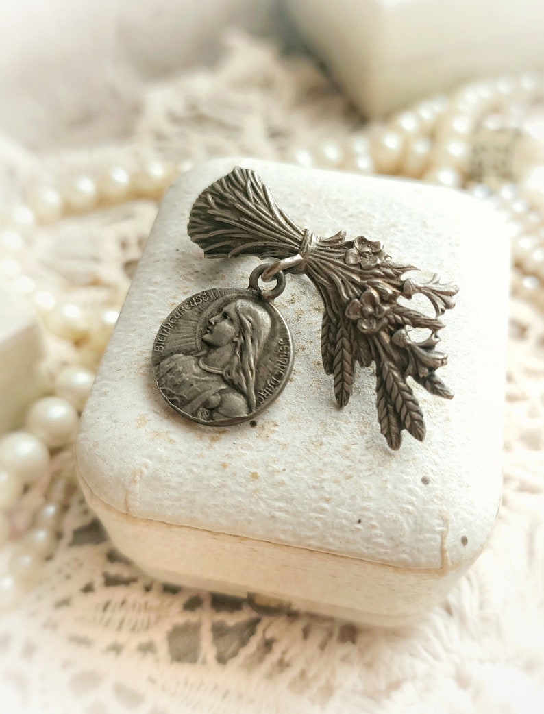 May include: A silver pendant featuring a religious medal with the image of a woman and the text "BEAUFIGURISE" and "SACI MARIE". The pendant is attached to a silver charm shaped like a bouquet of wheat.