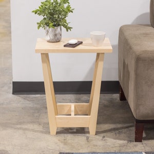 A side table placed in a living room with a plant and coffee mug sat on the table top. The side table is made from light maple hardwood and is crafted in Scandinavian minimalist modern design.