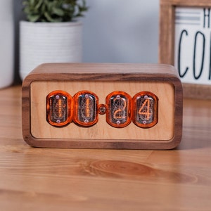 Clock sits on counter table made from solid walnut with a contrasting maple face. The design uses retro Nixie tubes for a warm orange glow.