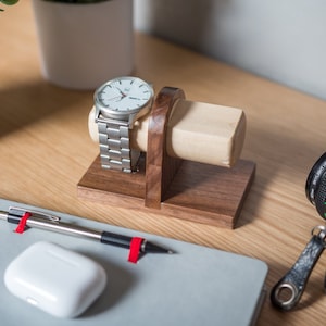 Two watches rest on a smooth maple dowel which rests on a strong walnut base. Set on a desk with notebook and camera. Light and warm wood contrasts well with mechanical metal watch.