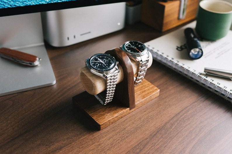 May include: Two silver wristwatches with black faces are displayed on a wooden watch stand. The stand is made of dark wood and has two slots for the watches. The watches are on a wooden desk.