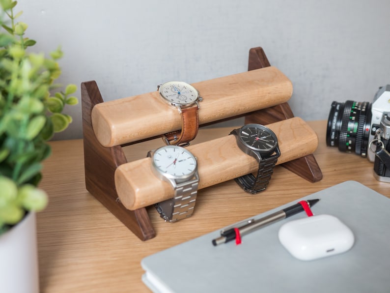 Three watches rest on a smooth maple dowel which rests on a strong walnut base. Set on a desk with notebook and camera.