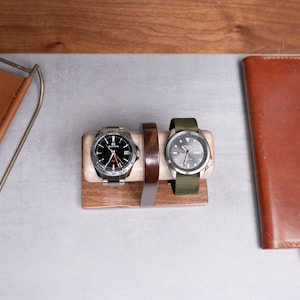 May include: Two wristwatches on a wooden watch stand. The watch on the left has a black face with silver hands and a silver band. The watch on the right has a silver face with black hands and a green fabric band.