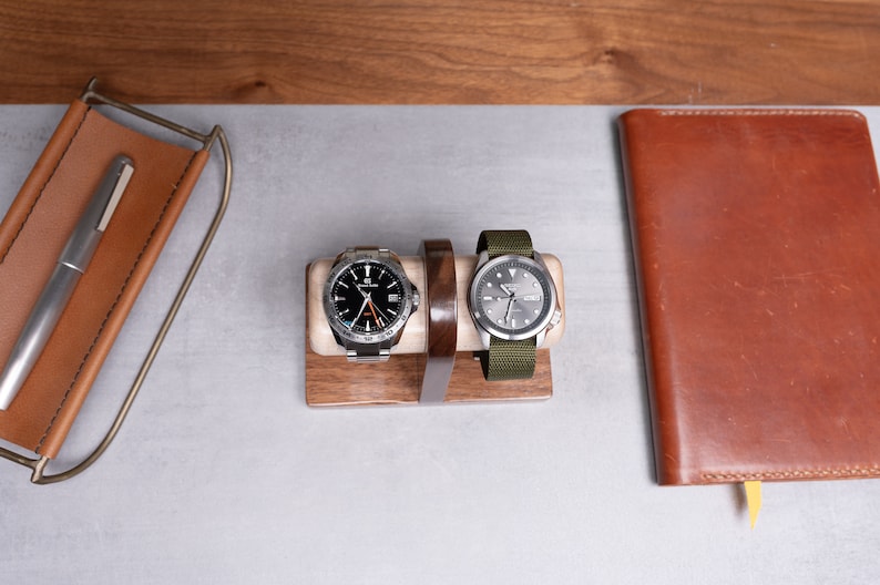May include: Two wristwatches on a wooden watch stand. The watch on the left has a black face with silver hands and a silver band. The watch on the right has a silver face with black hands and a green fabric band.