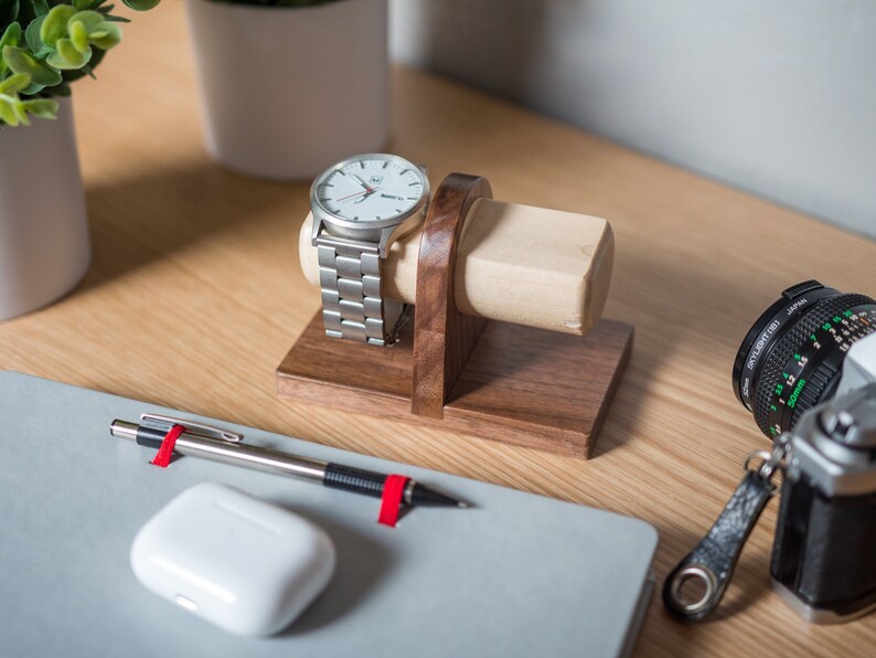 Two watches rest on a smooth maple dowel which rests on a strong walnut base. Set on a desk with notebook and camera. Light and warm wood contrasts well with mechanical metal watch.