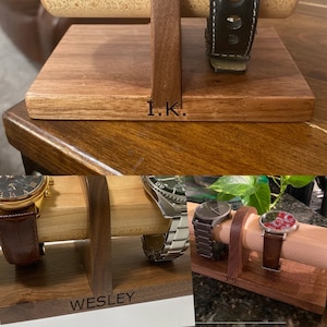 May include: Wooden watch stand with three slots for watches. The stand is made of dark wood and has a light wood top. The stand is engraved with the names "Wesley", "J.S.", and "I.K.".