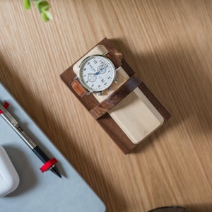 Two watches rest on a smooth maple dowel which rests on a strong walnut base. Set on a desk with notebook and camera. Light and warm wood contrasts well with mechanical metal watch.