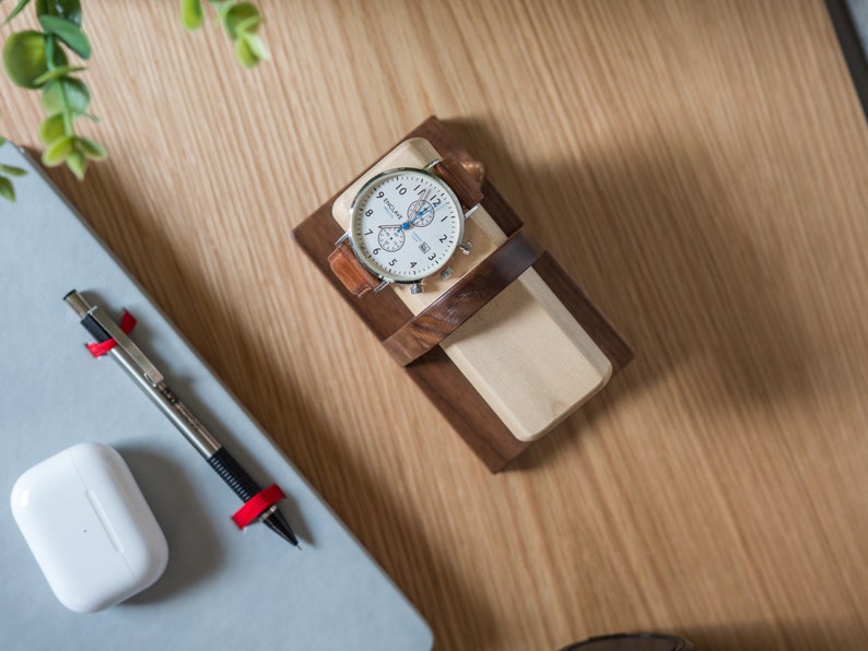 Two watches rest on a smooth maple dowel which rests on a strong walnut base. Set on a desk with notebook and camera. Light and warm wood contrasts well with mechanical metal watch.