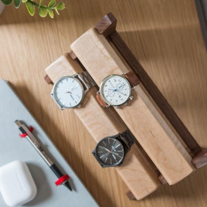 Three watches rest on a smooth maple dowel which rests on a strong walnut base. Set on a desk with notebook and camera.