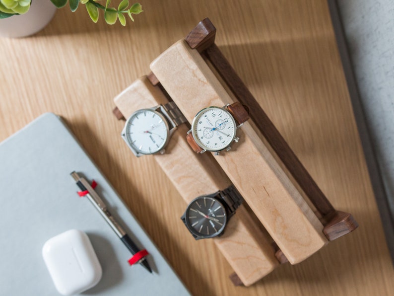 Three watches rest on a smooth maple dowel which rests on a strong walnut base. Set on a desk with notebook and camera.