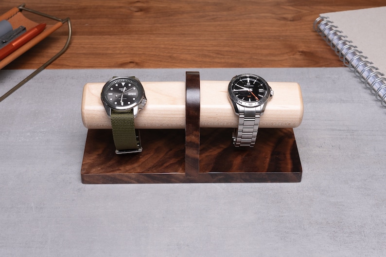 May include: Two wristwatches with different bands are displayed on a wooden watch stand. The stand is made of maple and walnut wood. The watch stand is on a gray surface.