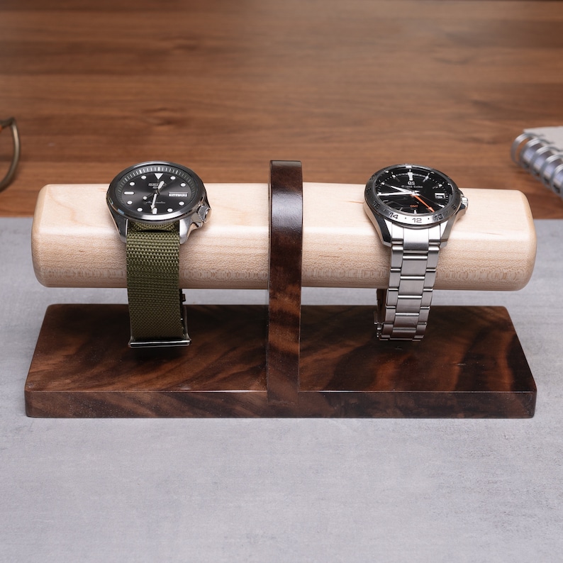May include: A wooden watch stand with two watch slots. The stand is made of dark wood with a light wood cylinder in the center. Two watches are displayed on the stand, one with a green strap and the other with a silver metal strap.
