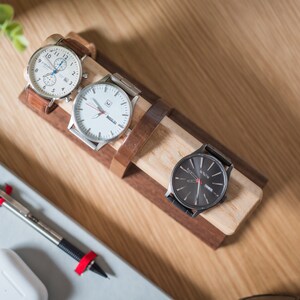 Watch display holder rests on a table, made of a long, wooden block base with one round dowel sticking out horizontally, enough space to hold four watches.