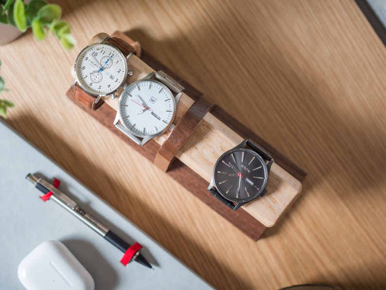 Watch display holder rests on a table, made of a long, wooden block base with one round dowel sticking out horizontally, enough space to hold four watches.