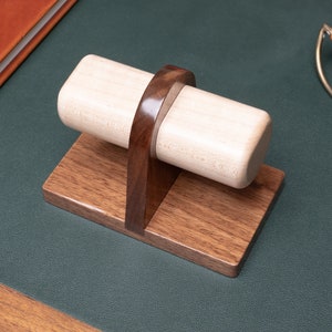 May include: A wooden watch winder with a light brown base and a dark brown stand. The winder is made of light wood and has a cylindrical shape.