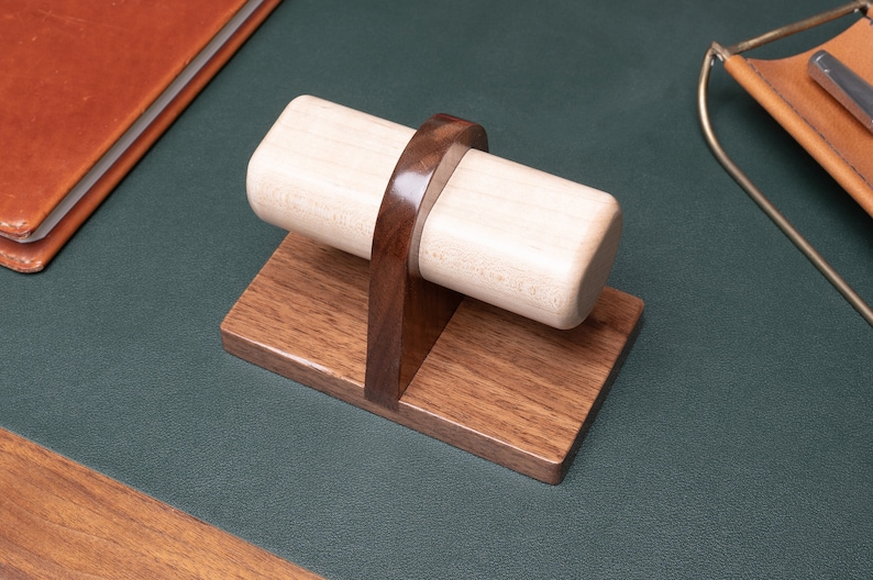 May include: A wooden watch winder with a light brown base and a dark brown stand. The winder is made of light wood and has a cylindrical shape.