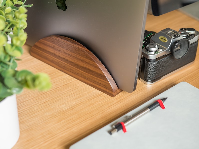 May include: A wooden laptop stand with a curved design, a silver camera, and a white notepad with a black pen on a wooden desk.