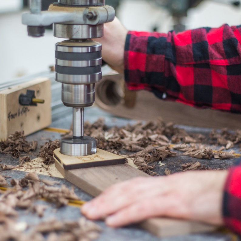 May include: A close-up of a drill press with a drill bit going through a piece of wood. The wood has the letters "IDT" engraved on it. There are wood shavings on the surface around the wood.