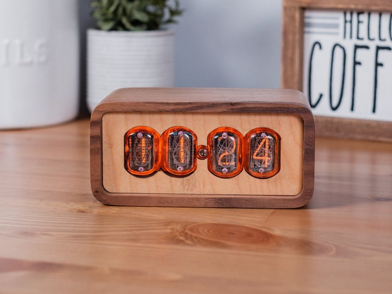 Clock sits on counter table made from solid walnut with a contrasting maple face. The design uses retro Nixie tubes for a warm orange glow.