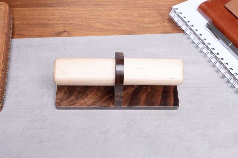 May include: A wooden pen holder with a dark brown base and a light wood top. The holder has a ring in the middle to hold the pen in place.