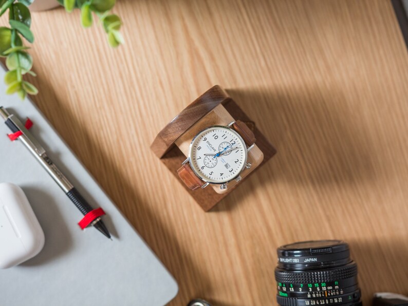 One watch rests on a smooth maple dowel which rests on a strong walnut base. Set on a desk with notebook and camera.