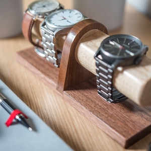Watch display holder rests on a table, made of a long, wooden block base with one round dowel sticking out horizontally, enough space to hold four watches.