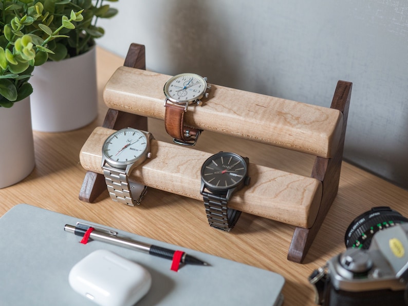 Three watches rest on a smooth maple dowel which rests on a strong walnut base. Set on a desk with notebook and camera. Warm wood nicely contrasts the mechanical metal watch.