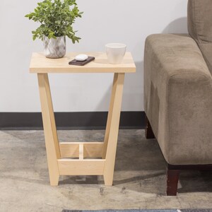 A side table placed in a living room with a plant and coffee mug sat on the table top. The side table is made from light maple hardwood and is crafted in Scandinavian minimalist modern design.