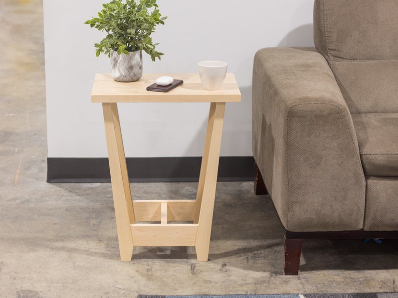 A side table placed in a living room with a plant and coffee mug sat on the table top. The side table is made from light maple hardwood and is crafted in Scandinavian minimalist modern design.